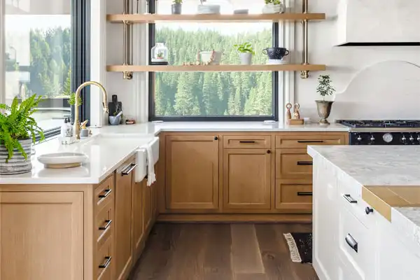 Modern kitchen with light wood cabinets and white countertops, overlooking greenery. Open shelves hold small plants and dishes. A corner sink with a gold faucet adds elegance, combining style with practicality—just in case you need quick plumbing repair from a plumber nearby.
