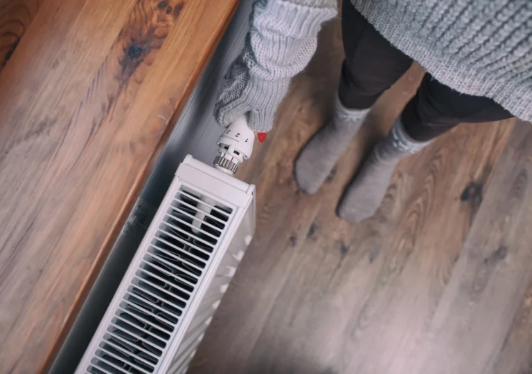 Person wearing a gray sweater and gloves adjusts the thermostat on a radiator. They are standing on a wooden floor, and the view is from above.
