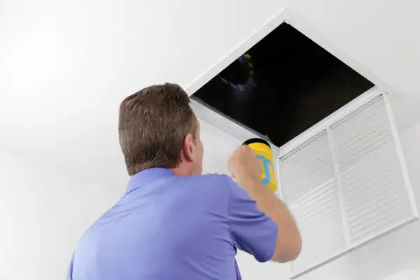 A man in a blue shirt shines a flashlight into a ceiling access panel, inspecting the dark space inside.