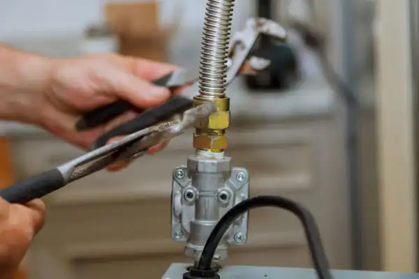 Person using pliers and wrench to tighten a valve on a flexible metal hose connected to machinery.