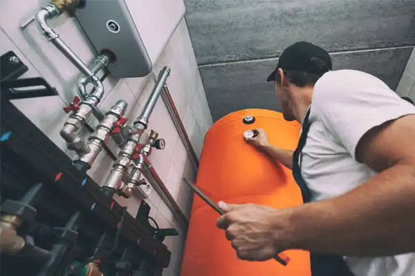 Worker in a cap and overalls, adjusting valves on a large orange industrial tank in a mechanical room.