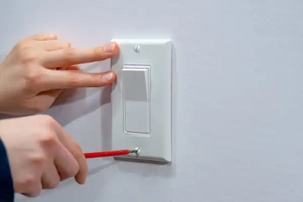 Hands adjusting a white light switch cover on a wall using a red screwdriver.