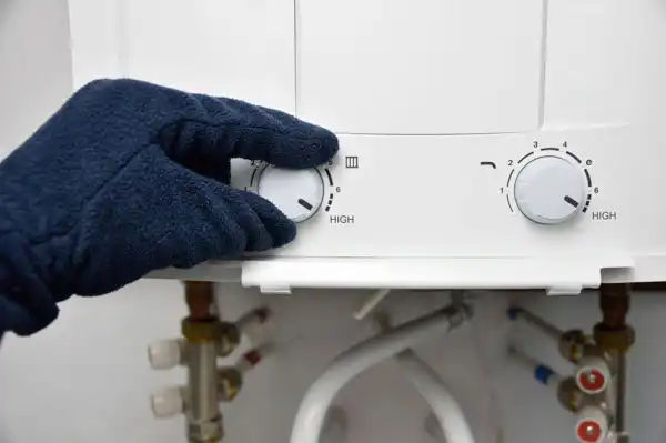 A gloved hand adjusts the temperature knob on a water heater, showing settings labeled "High.